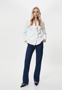 Blazer bianco doppiopetto in tessuto testurizzato, con due tasche frontali. Abbinato a jeans svasati blu scuro e tacchi a punta neri.