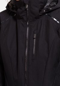 Svart isolerad jacka med hög krage, blanka detaljer, dragkedjefickor och en texturerad tygdesign. Har en vattentät finish.