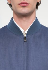 Μπλε μπουφάν με ριμπ κολάρο, μπροστινό φερμουάρ και ομαλή υφή υφάσματος. Από κάτω διακρίνεται μερικώς ένα λευκό πουκάμισο.