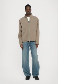 Chaqueta taupe con cremallera y cuello alto, que presenta dos bolsillos frontales y herrajes plateados. Combinada con unos vaqueros de denim azul de corte ancho y zapatos negros.