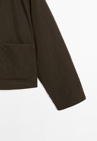 Veste en tissu texturé marron avec une poche frontale unique et des manches longues, taillée avec un ourlet légèrement incliné.