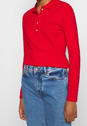 Personne portant un polo rouge à manches longues rentré dans un jean bleu taille haute avec une fermeture à boutons et à glissière.