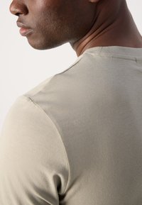 Gai olīvu zaļas krāsas T-krekls ar gludu adījuma faktūru un apaļu izgriezumu, kurā redzami smalki iestrādāti šuves lējumi uz pleca.