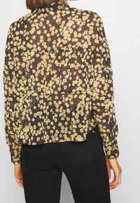 Blusa nera trasparente con motivo floreale giallo e rosso, maniche lunghe e vestibilità comoda. Il dettaglio della piega posteriore aggiunge texture.