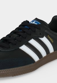 Sort Adidas Samba sneaker med hvide striber, brun sål, sorte snørebånd, blå logo på tungen og gylden tekst på siden.