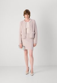 Lichtroze textuurjasje met voorkruiszakken, gecombineerd met een bijpassende getailleerde rok. Houdt een zilveren clutch vast en draagt naakte two-tone hakken.