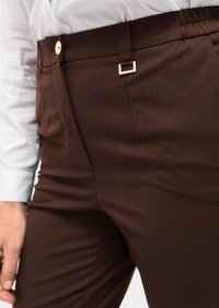 Bruine op maat gemaakte broeken met een gladde textuur, uitgerust met een vierkante lus in goudkleur en een voorknop. Draagt met een lichtblauw shirt.