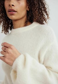 Kreminė, tekstūruota megzta palaidinė su apvaliu iškirpimu ir plačiomis, švelniai siaurėjančiomis rankovėmis. Lygus audinys su subtiliomis spalvų variacijomis.