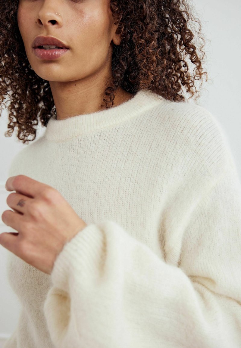 Kreminė, tekstūruota megzta palaidinė su apvaliu iškirpimu ir plačiomis, švelniai siaurėjančiomis rankovėmis. Lygus audinys su subtiliomis spalvų variacijomis.