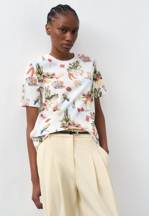 Woman wearing white short-sleeve shirt with colorful desert and bird illustrations, tucked into high-waisted beige trousers with a black belt.