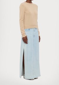 Personne portant un pull à manches longues beige avec des poignets à volants, une maxi jupe en jean bleu clair avec une fente latérale et des chaussures argentées.