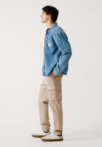 Lichtblauwe spijkerjas met een borstzak; beige cargobroek met twee zijzakken; witte sneakers met zwarte accenten en bruine zolen.