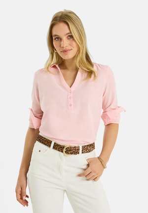 Mujer rubia con blusa rosa abotonada y mangas remangadas, pantalones blancos, cinturón de estampado de leopardo y pulsera dorada en la muñeca derecha.