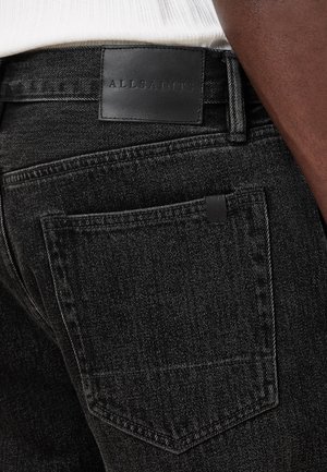 Nahaufnahme einer schwarzen Denim-Jeanstasche mit schwarzem Lederetikett der Marke „AllSaints“ am Bund und einem dunkelhäutigen Arm daneben.