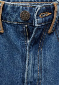 Gros plan de la taille d'un jean en denim bleu avec une braguette partiellement zippée, un bouton métal et des coutures visibles sur les passants de ceinture et les coutures.