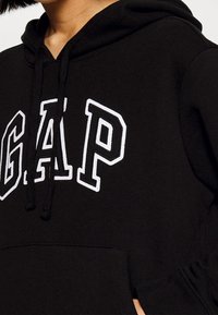 Sweat à capuche noir avec logo "GAP" blanc, doté d'une capuche avec cordon de serrage et d'une poche frontale. Confectionné en tissu doux, avec une texture côtelée sur les poignets.