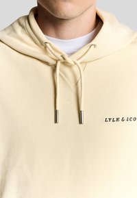 Cremefarvet hættetrøje med snorelukning, sølvfarvede aglets, og et broderet "LYLE & SCO" logo på venstre bryst. Blød tekstur.