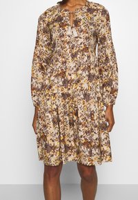 Kukallinen mekko beige, ruskea ja keltainen sävyissä. Sisältää avonaisen kaula-aukon nauhalla ja tupsuilla, sekä pitkät, kerätyt hihat.