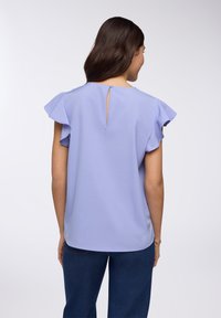 Blusa lilla con maniche a ruffle, scollo rotondo e dettaglio a chiave sulla schiena, realizzata in tessuto leggero. Abbinata a pantaloni scuri.