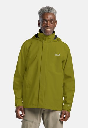 Smiling man with gray hair wearing a green Jack Wolfskin waterproof jacket and beige cargo pants against a plain background.