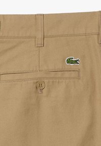 I pantaloni in cotone beige presentano una tasca posteriore con un logo di coccodrillo verde e una chiusura con bottone marrone. Il tessuto ha una finitura liscia e strutturata.