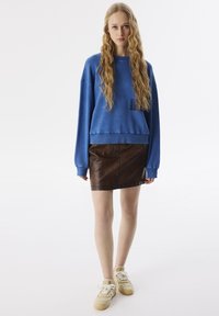 Blauwe sweatshirt met lange mouwen, gecombineerd met een bruine leren minirok. Model draagt beige sneakers met textuurelementen.