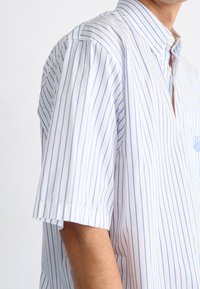 Lyhythihainen nappipaita valkoisena, jossa siniset pystysuorat raidat, teräväkaulus ja rintatasku. Valmistettu kevyestä kankaasta.