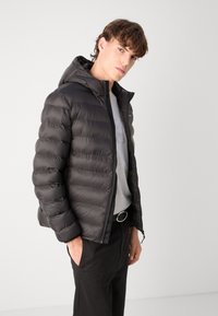 Giubbotto a pistone nero con imbottitura orizzontale, cappuccio e zip frontale. Indossato sopra una maglietta grigia, abbinato a pantaloni neri e una fibbia circolare per la cintura.