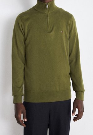 Homme portant un pull zippé vert olive avec un petit logo sur la poitrine et un pantalon noir sur fond uni.