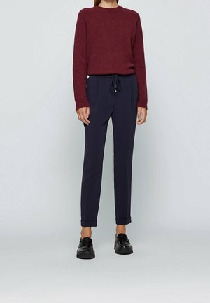 Vrouw draagt een bordeauxrode trui met lange mouwen, marineblauwe broek met trekkoord en omgeslagen pijpen, en zwarte loafers, staand tegen een effen achtergrond.