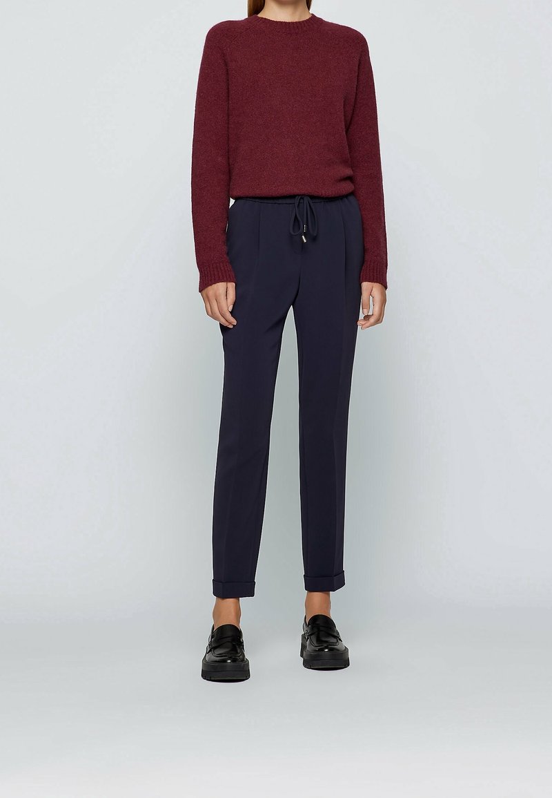 Femme portant un pull à manches longues bordeaux, un pantalon bleu marine à cordon de serrage avec ourlets retroussés, et des mocassins noirs, debout devant un fond uni.