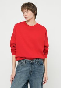 Oseba s kratkimi rjavimi lasmi, oblečena v rdečo dolg rokav sweatshirt, vsajen v hlače z visokim pasom v modri barvi, stoji ob navadnem belem ozadju.