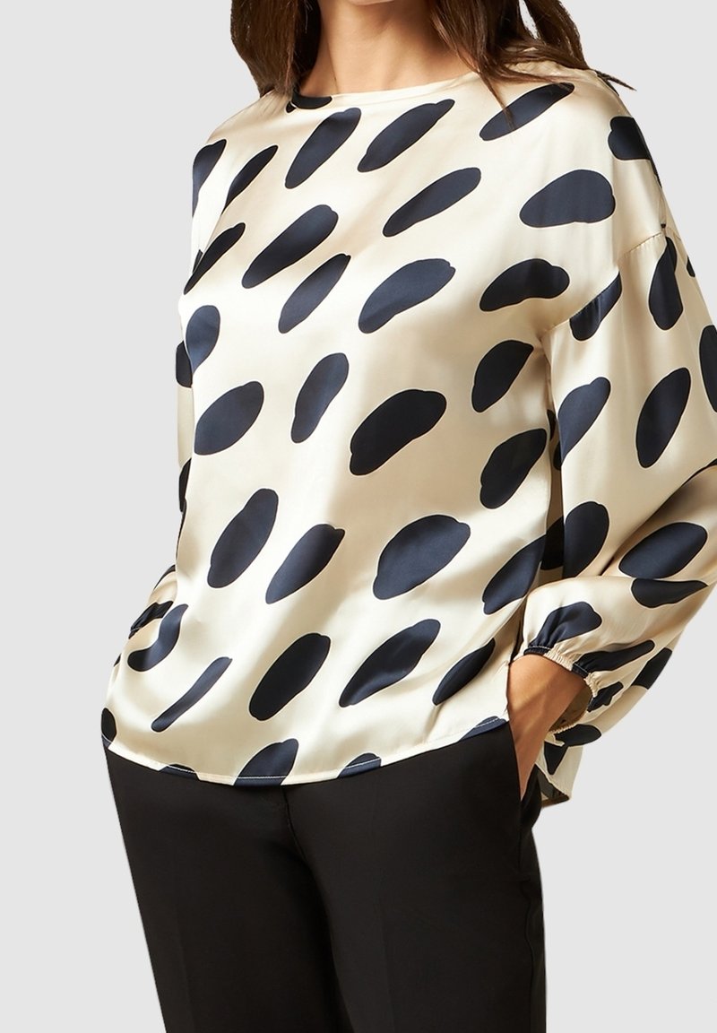 Femme portant une blouse en satin crème avec de grandes taches noires irrégulières, manches longues, et un pantalon noir, mains dans les poches, sur un fond uni.