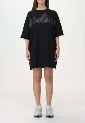 Abito t-shirt nero oversize con maniche corte e testo in strass. Tessuto morbido. Abbinato a scarpe sportive grigio chiaro.