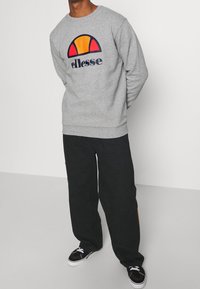 Sweat-shirt gris avec un logo coloré comprenant des arcs orange et rouge, accompagné d'un pantalon noir ample et de baskets noires avec des accents blancs.