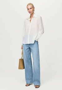 Weißes, langärmliges Hemd mit Knopfleiste und bestickten Details, kombiniert mit hellblauen, weit geschnittenen Jeans und einer braunen Tragetasche. Schwarze Sandalen.