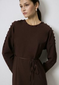 Smeđa haljina dugih rukava s okruglim izrezom, ukrašena pom-pom detaljima duž rukava i vezanom strukom, izrađena od glatkog materijala.