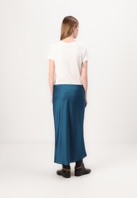 Lunga gonna in satin teal con una texture liscia, abbinata a una t-shirt bianca a maniche corte e stivali neri alla caviglia con un accento a catena.
