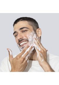 Środek czyszczący na twarzy uśmiechniętego mężczyzny. Ma krótke ciemne włosy, jasną skórę i nosi beżowy t-shirt. Pianka jest na jego rękach.
