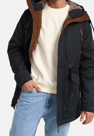 Homme portant une veste à capuche noire avec une doublure marron sur un pull blanc texturé et un jean bleu clair, une main dans la poche.