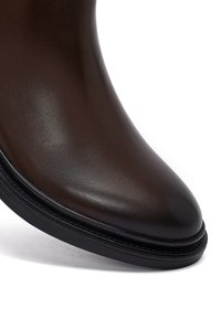 Primo piano di una scarpa in pelle marrone scuro con punta tonda e suola nera su sfondo bianco.