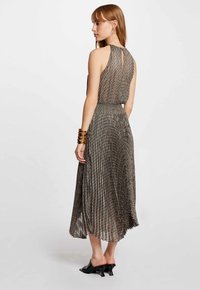 Plissiertes Midi-Kleid mit einem Schlüsselrücken, das ein strukturiertes Schwarz-Gold-Muster aufweist. Begleitet von einem massiven goldenen Armband und Absatzschuhen.