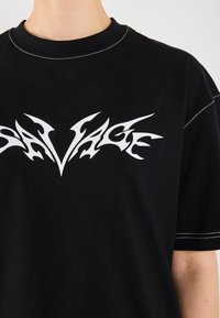 Black cotton t-shirt with a round neckline and white "SAVAGE" graphic print. Features contrast stitching on the sleeves.