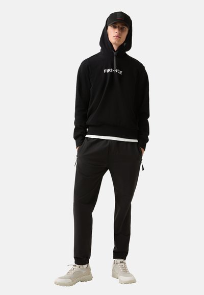 Jonge man in een zwarte hoodie met de tekst "FIRE + ICE", een zwarte pet, zwarte broek en witte sneakers, staande met zijn handen in zijn zakken.