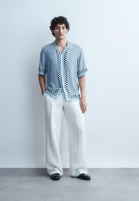 Jonge man met krullend haar, gekleed in een lichtblauw shirt met korte mouwen en patroon, witte broek met wijd uitlopende pijpen en zwarte schoenen, staande tegen een effen muur.