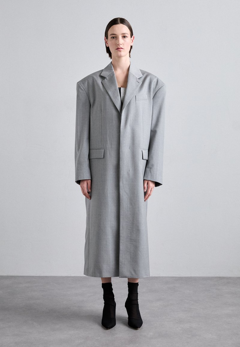 Blazer abu-abu panjang terbuat dari kain tenun, memiliki bahu oversized, kantong tempel, dan siluet lurus. Dipakai di atas atasan hitam dan sepatu bot hak tinggi.