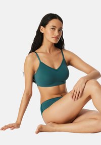 Kobieta o długich ciemnych włosach siedząca z nogami skrzyżowanymi, ubrana w morski biustonosz i dopasowane majtki na jednolitym białym tle.