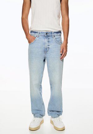 Persoon draagt lichtblauwe versleten rechte spijkerbroek, witte sneakers met beige zolen en een mouwloos wit geribd topje.