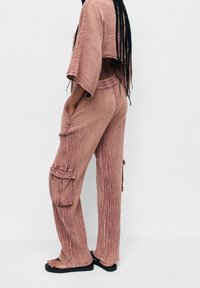 Personne aux longs cheveux tressés portant un pantalon cargo rose ample et texturé avec un haut court assorti, debout en sandales noires sur fond blanc.