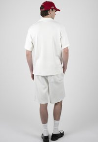 Jeune homme portant une casquette rouge, une chemise blanche à manches courtes, un short blanc, des chaussettes blanches et des chaussures noires, debout de dos sur un fond uni.